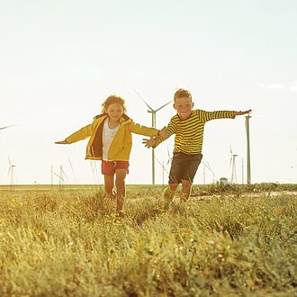 Zwei Kinder toben mit ausgestreckten Armen vor Windenergieanlagen im Sommersonnenlicht